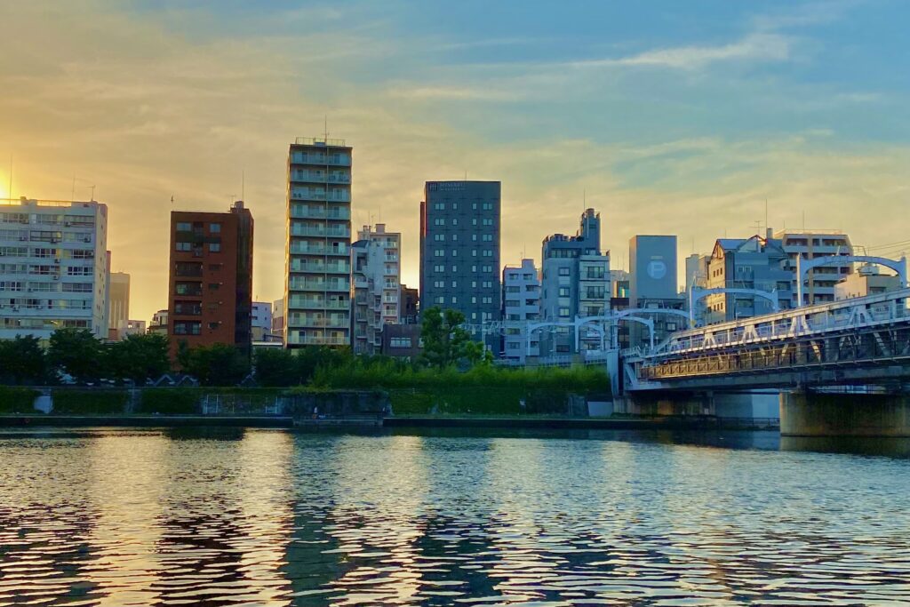 墨田区側の川岸の歩道からの風景です。浅草の町並みと川面を照らす夕焼けのオレンジと空と水面の青色が織りなすグラデーションが印象的(撮影：TOE LIBRARY)